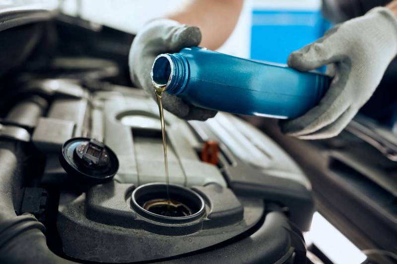 A car with an open bonnet has its engine oil topped up from a blue bottle